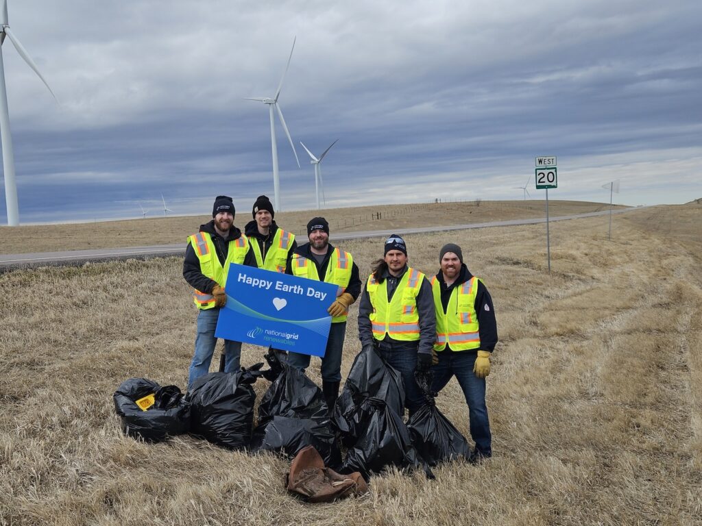 People with Happy Earth Day sign and trash bags next to highway with wind turbines in background